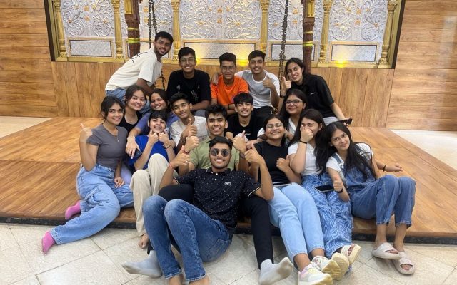 Gitam Junior College Students – Group Bonding Moment Group of cheerful college students posing together indoors on a wooden stage with traditional decor in the background.